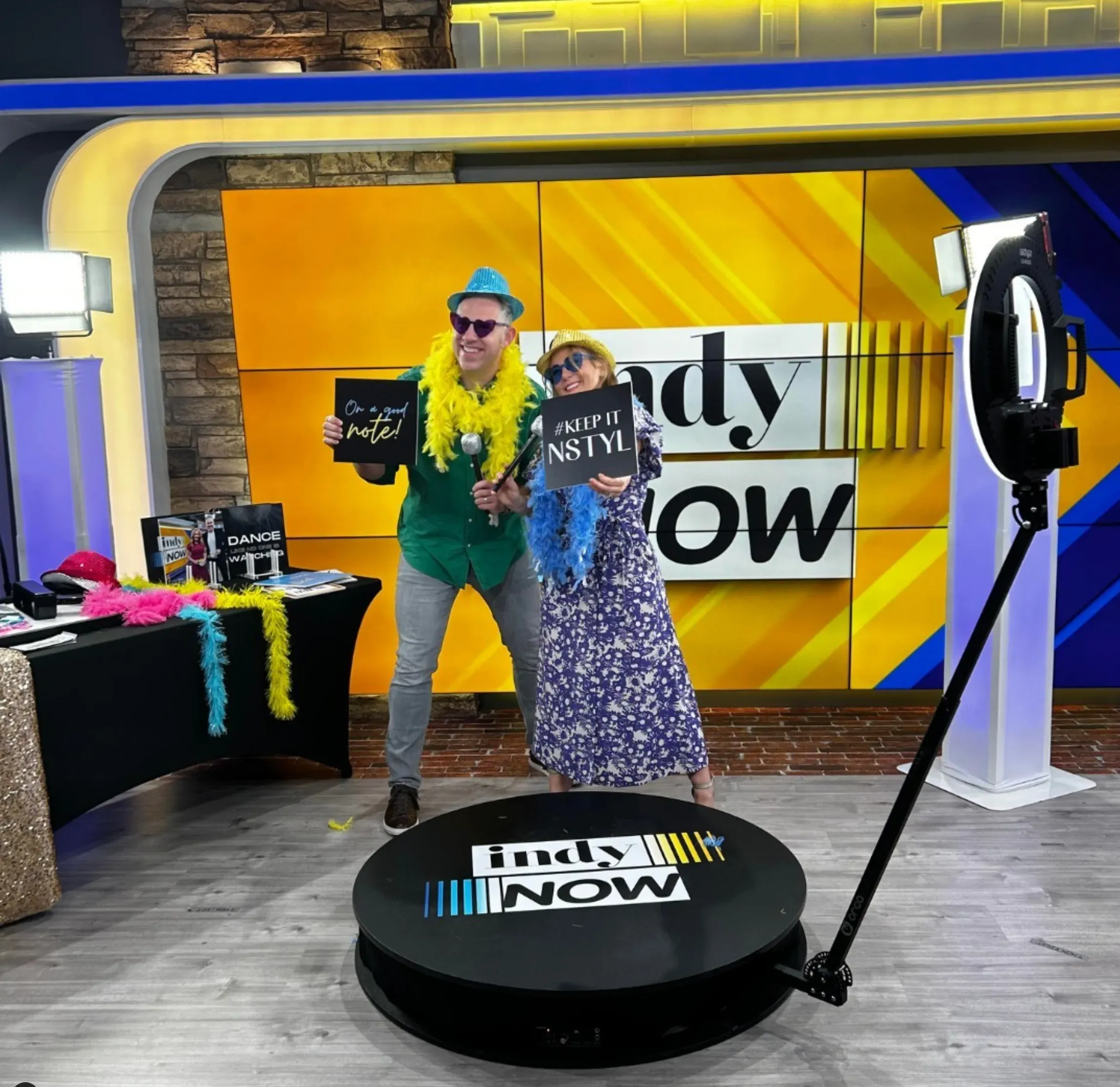 Two people pose on a branded 360° photo booth platform labeled 'Indy Now,' wearing colorful feather boas, sunglasses, and hats while holding signs that read 'On a good note!' and '#KEEP IT NSTYL.' They're in a lively TV studio set with bright yellow and blue graphics, a ring light, and a decorated table with props nearby.