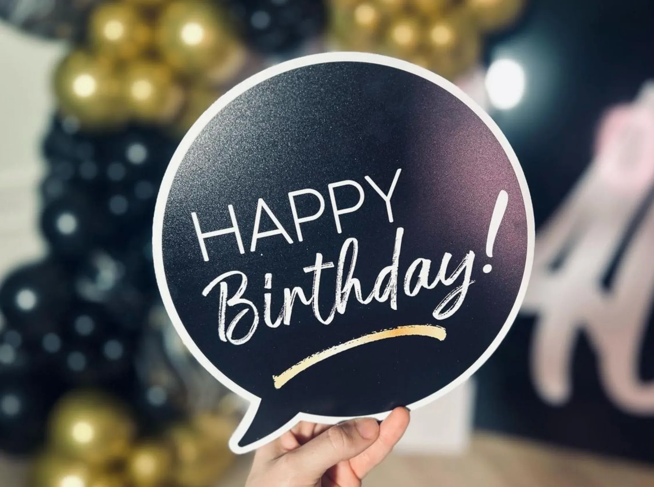 Close-up of a hand holding a black photo booth prop sign shaped like a speech bubble with the words “Happy Birthday!” in white and gold.