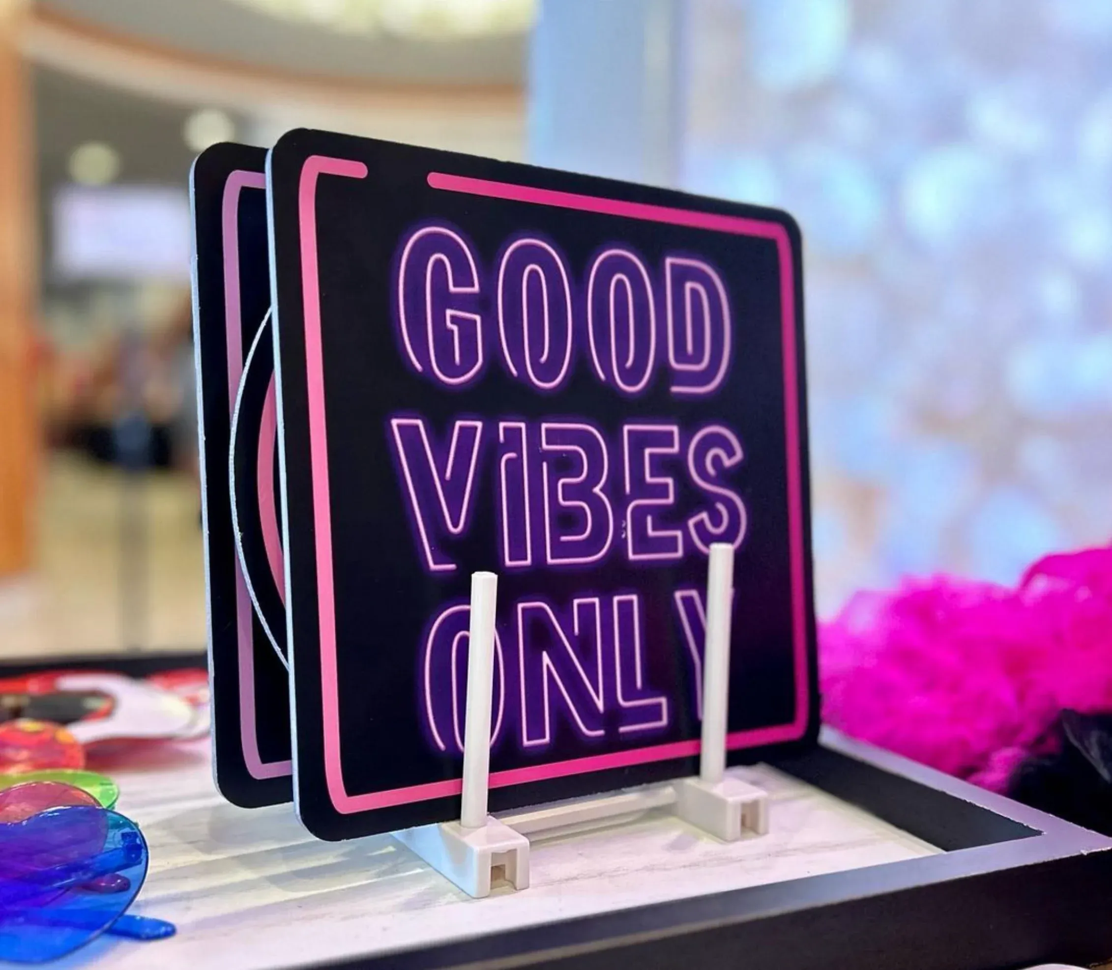 Photo booth prop sign that reads “GOOD VIBES ONLY” in neon-style lettering, displayed on a tabletop with colorful accessories and a pink feather boa nearby.
