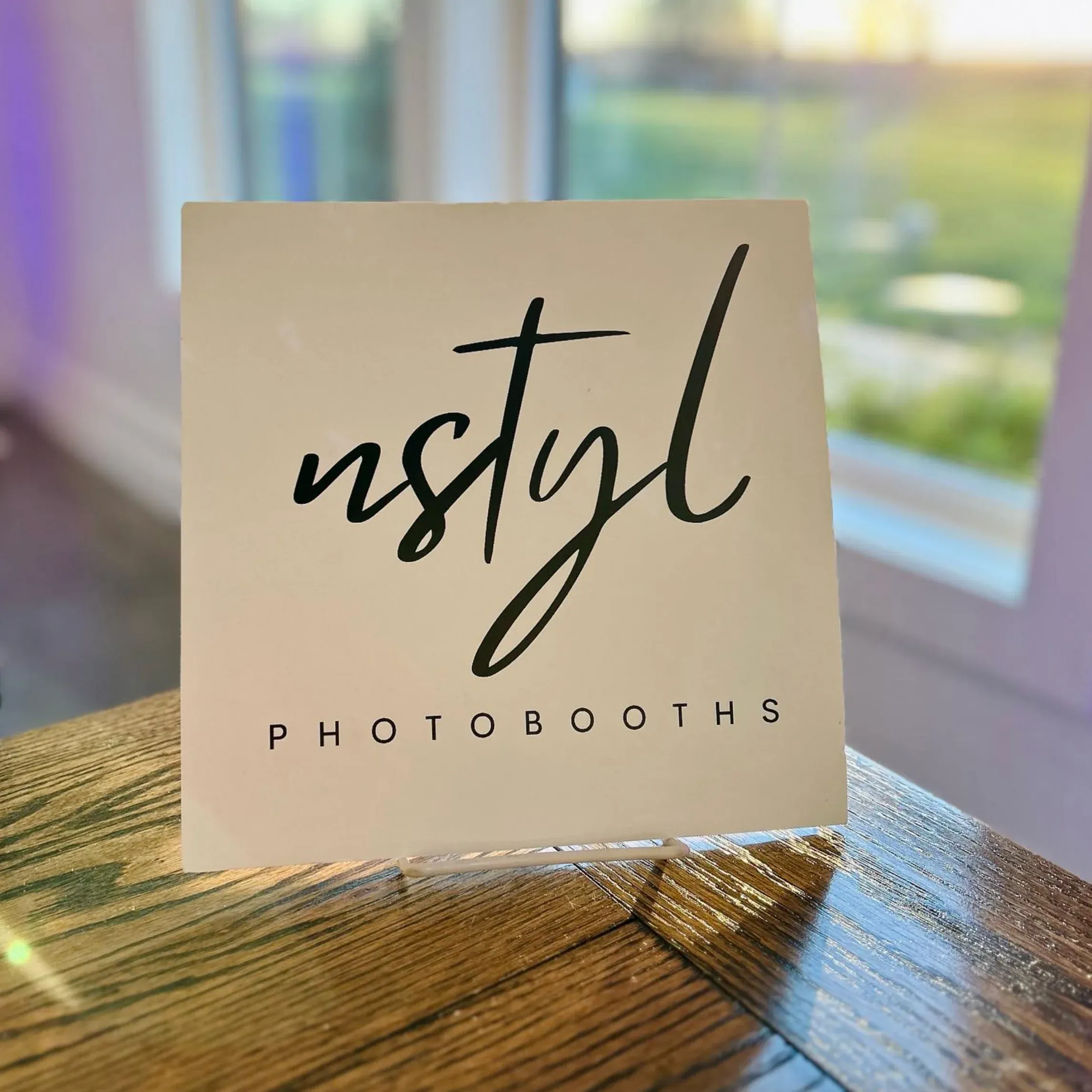 Small tabletop display sign with the Nstyl Photobooths logo in black script on a cream background, sitting on a wooden surface with soft daylight in the background.